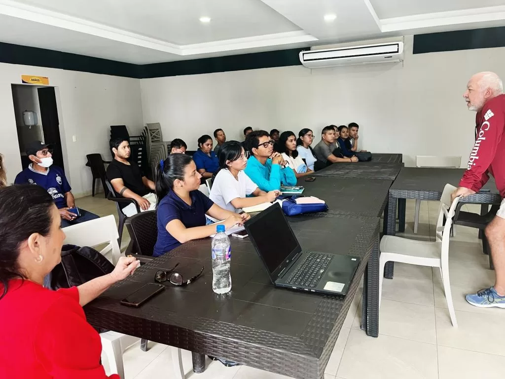 Sala de aula com instrutor à frente e turma de jovens atentos, mesas em U, notebooks e cadernos; ambiente de capacitação técnica em aquicultura.