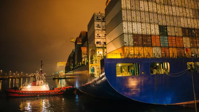 Contêineres parados em porto à noite, com navio de carga