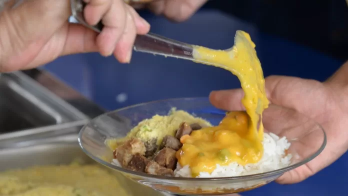 Merenda escolar com filé de camarão em Sergipe Prato de merenda escolar com arroz, farofa, carne e molho de camarão sendo servido