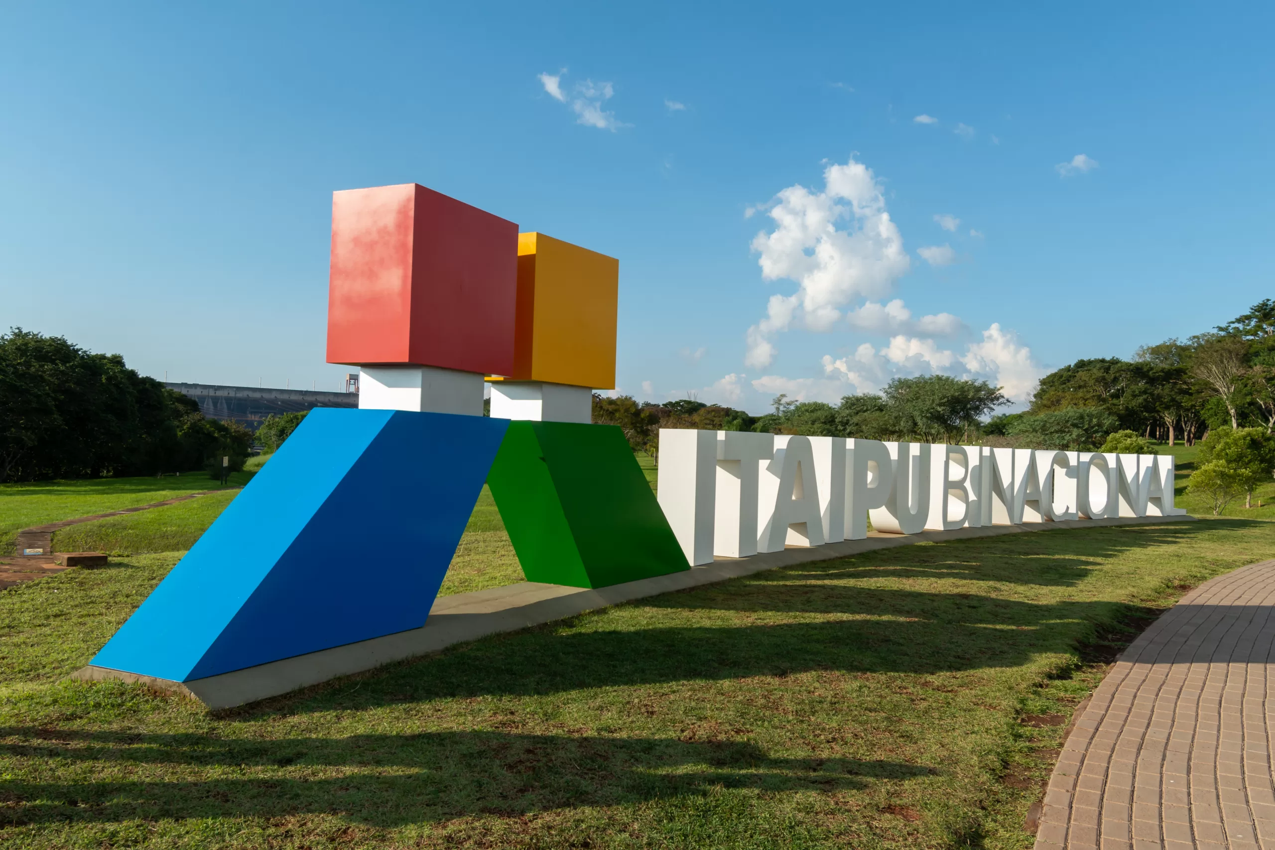 Letreiro turístico colorido com o nome Itaipu Binacional sobre gramado verde em dia ensolarado.