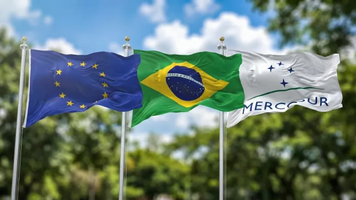 Bandeiras da União Europeia, do Brasil e do Mercosul hasteadas lado a lado ao ar livre, simbolizando o acordo comercial.
