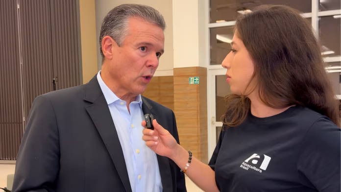 Foto do Ministro André de Paula, que deixa a Pesca para assumir o Ministério da Agricultura (MAPA), sendo entrevistado pela repórter da Aquaculture Brasil. Ele usa terno preto e camisa azul claro; ela segura um pequeno microfone. O fundo mostra um centro de convenções.