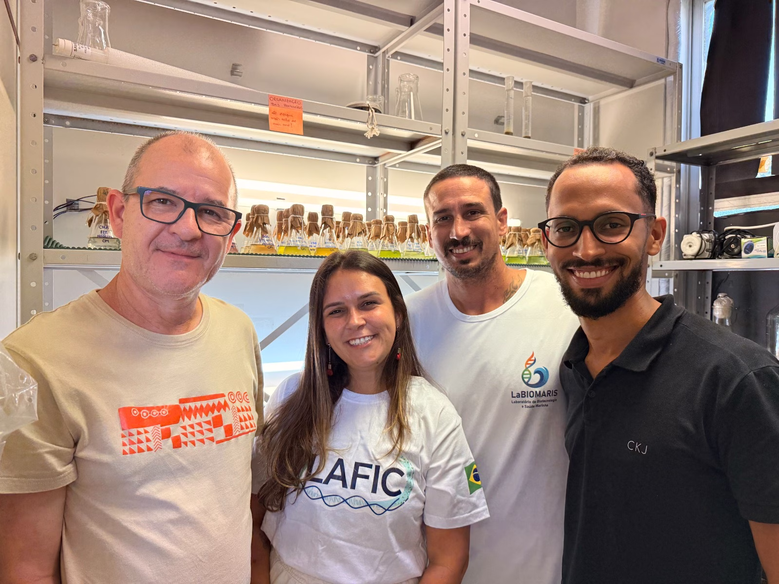 Quatro pesquisadores sorrindo e posando para foto em um laboratório, em frente a prateleiras com frascos contendo líquidos de diferentes cores.