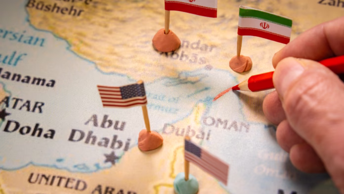 Mão desenhando uma linha vermelha com lápis no Estreito de Ormuz em um mapa com bandeiras dos EUA e do Irã.