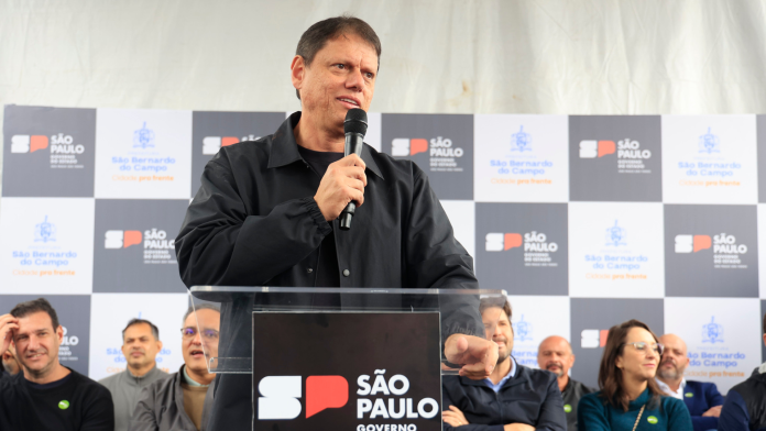 Governador de São Paulo, Tarcísio de Freitas, discursando em um microfone durante evento oficial com painel de fundo institucional do Governo do Estado.