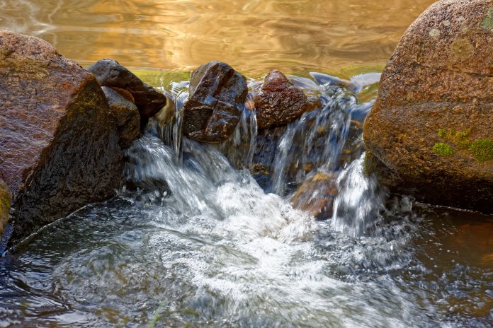 Uma pequena cascata de água em um córrego de montanha fluindo sobre rochas grandes. Água branca espumante cai entre pedras arredondadas e texturizadas. Luz dourada reflete no riacho acima da queda. Uma rocha no lado direito tem musgo verde.