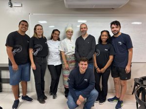 Grupo de pesquisadores, professores e estudantes, alguns com uniformes de gastronomia e outros com camisetas de laboratório, posando juntos para foto em uma sala com quadro branco.