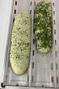 Duas massas de pão cruas e alongadas, salpicadas com pequenos pedaços verdes, dispostas sobre uma fôrma metálica perfurada de panificação.